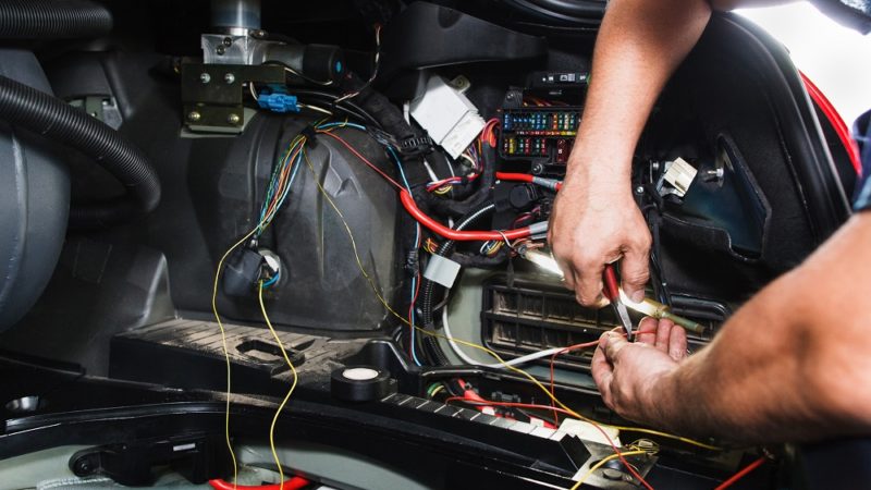 Electrician works with electric block in car. Close-up of automobile inside under raised hood. Service man hands working with cables of auto